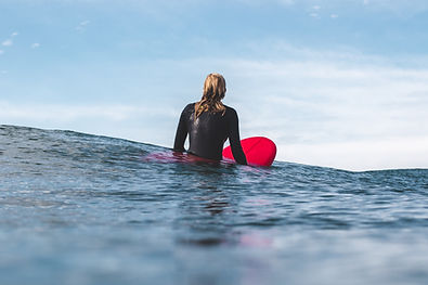 Woman in a black wetsuit sitting on a red surfboard in the ocean, gazing at the horizon under a soft blue sky Woman in a black wetsuit sitting on a red surfboard in the ocean, gazing at the horizon under a soft blue sky