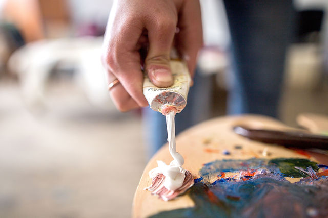 Close-up of a hand squeezing white paint from a tube onto a wooden palette smeared with blue, green and orange paint Close-up of a hand squeezing white paint from a tube onto a wooden palette smeared with blue, green and orange paint