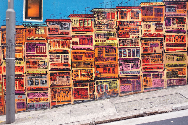 Vibrant street mural of stacked apartment buildings painted in orange, red, pink and purple along a sloping sidewalk against a blue wall