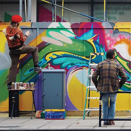 Street artist in a red beret spray-painting a bold, colorful abstract mural on a wall as a man on a ladder watches