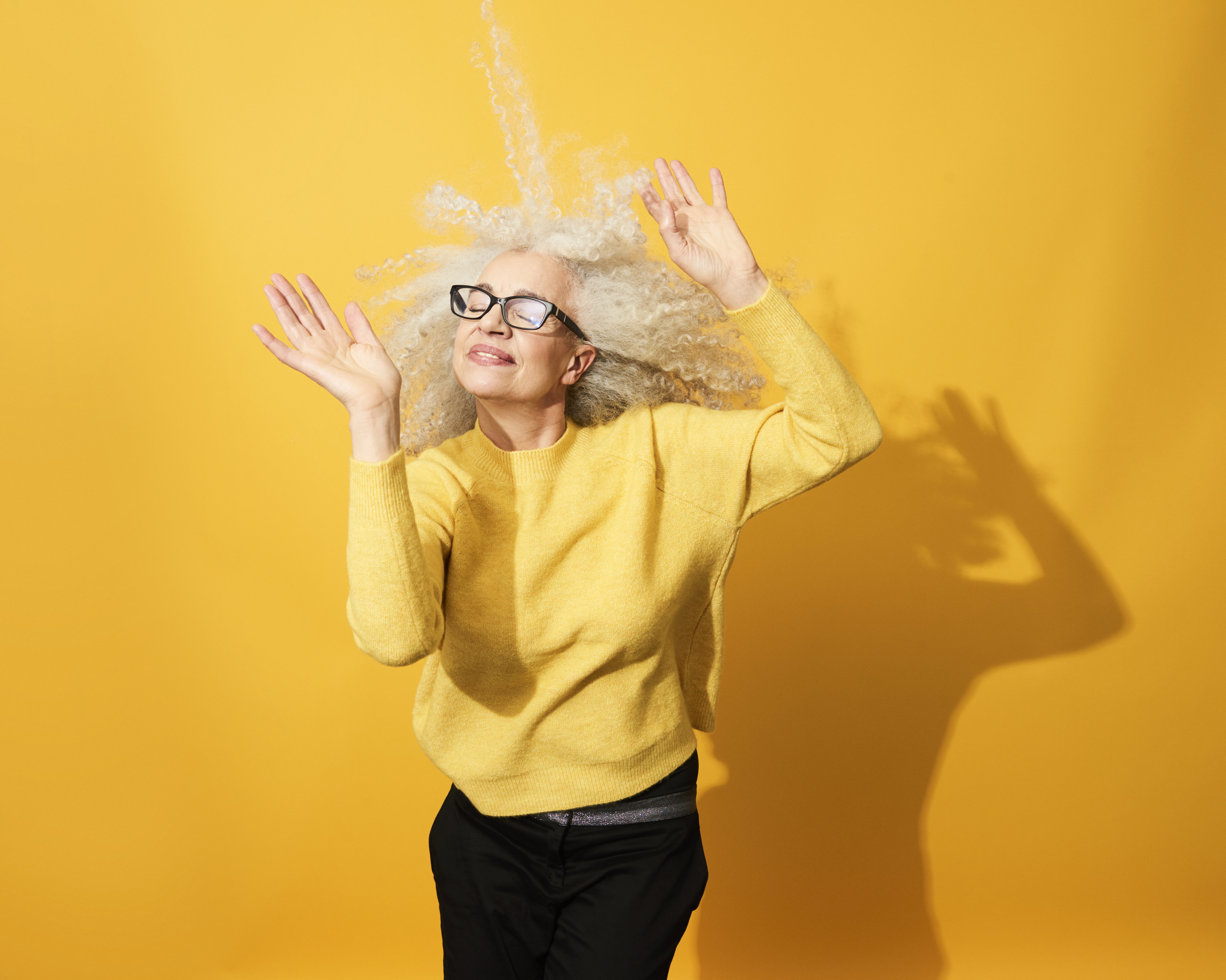 Older woman with wild silver curls and glasses dancing with eyes closed and hands raised in a yellow sweater against a bright yellow wall Older woman with wild silver curls and glasses dancing with eyes closed and hands raised in a yellow sweater against a bright yellow wall