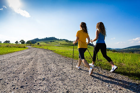 Two women Nordic walking with poles along a gravel country road through green hills under a bright blue sky Two women Nordic walking with poles along a gravel country road through green hills under a bright blue sky