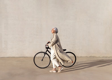 Older woman with silver hair in a long beige coat walking her bicycle in the sunshine against a pale wall, face tilted toward the light Older woman with silver hair in a long beige coat walking her bicycle in the sunshine against a pale wall, face tilted toward the light