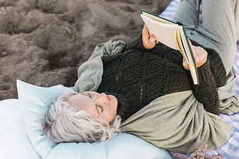 Older woman with silver hair lying on a pillow on the sand, wrapped in a cozy knit sweater and reading a book Older woman with silver hair lying on a pillow on the sand, wrapped in a cozy knit sweater and reading a book