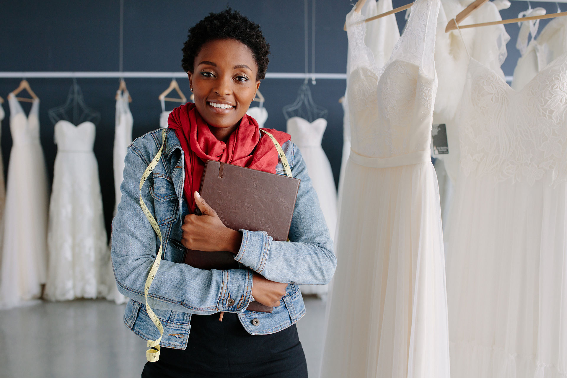 Smiling designer with short natural hair, a denim jacket and a red scarf, holding a leather notebook with a tape measure around her neck, in a bridal studio lined with white gowns Smiling designer with short natural hair, a denim jacket and a red scarf, holding a leather notebook with a tape measure around her neck, in a bridal studio lined with white gowns