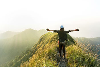 Hiker in a cap standing on a grassy mountain ridge with arms outstretched, facing hazy golden sunlight over rolling hills Hiker in a cap standing on a grassy mountain ridge with arms outstretched, facing hazy golden sunlight over rolling hills