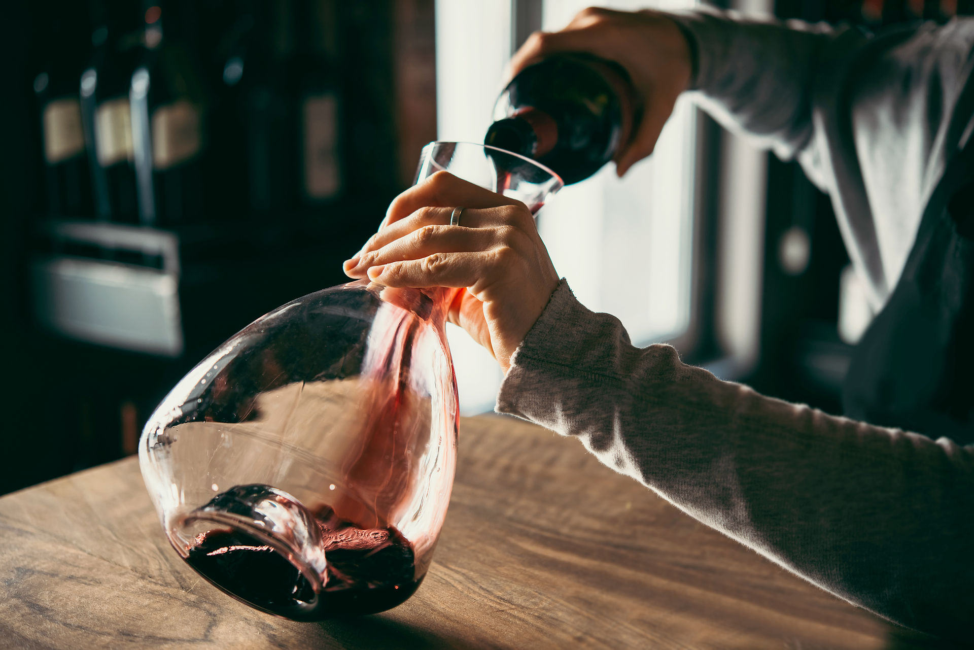 Hands pouring red wine from a bottle into a glass decanter on a wooden table, lit by soft window light Hands pouring red wine from a bottle into a glass decanter on a wooden table, lit by soft window light