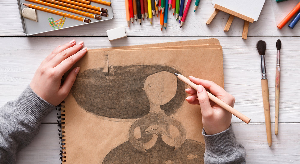 Overhead view of hands drawing a whimsical pencil sketch of a woman with flowing hair in a kraft-paper sketchbook, surrounded by colored pencils, paintbrushes and a small easel on a white wooden table Overhead view of hands drawing a whimsical pencil sketch of a woman with flowing hair in a kraft-paper sketchbook, surrounded by colored pencils, paintbrushes and a small easel on a white wooden table