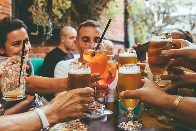 Group of friends raising glasses of beer and orange spritz cocktails in a toast at an outdoor bar table, with a smiling woman on the left