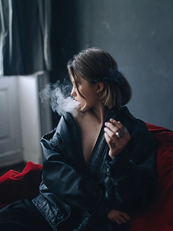 Young woman with a short bob in an oversized black leather jacket exhaling smoke while seated on a red couch in a dim room, looking toward the window