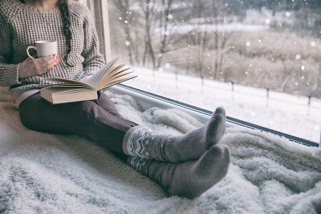 Woman in a knit sweater and wool socks sitting on a fluffy blanket by a window, holding a cup and an open book while snow falls outside Woman in a knit sweater and wool socks sitting on a fluffy blanket by a window, holding a cup and an open book while snow falls outside