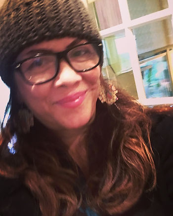 Smiling woman with long auburn wavy hair, black-framed glasses, a knit beanie and statement earrings taking a selfie indoors