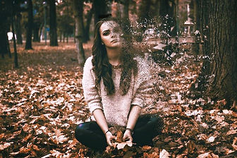 Sad young woman with long dark hair sitting cross-legged on fallen autumn leaves in a park, with part of her body dissolving into scattered particles Sad young woman with long dark hair sitting cross-legged on fallen autumn leaves in a park, with part of her body dissolving into scattered particles