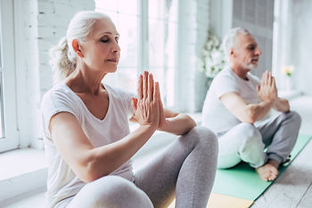 : Older woman and man sitting cross-legged on yoga mats with eyes closed and hands in prayer position, meditating in a bright white room : Older woman and man sitting cross-legged on yoga mats with eyes closed and hands in prayer position, meditating in a bright white room
