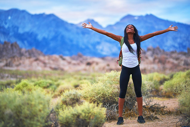 Joyful woman with long dark hair and a backpack standing with arms spread wide in a desert landscape of green shrubs, with blue mountains behind her