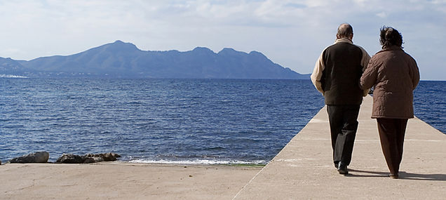 Older couple walking side by side along a stone pier toward the deep blue sea, with mountains on the horizon Older couple walking side by side along a stone pier toward the deep blue sea, with mountains on the horizon