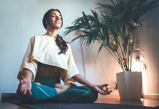 Smiling woman sitting cross-legged on a yoga mat with eyes closed and hands resting on her knees in meditation, beside a potted palm in a softly lit room Smiling woman sitting cross-legged on a yoga mat with eyes closed and hands resting on her knees in meditation, beside a potted palm in a softly lit room