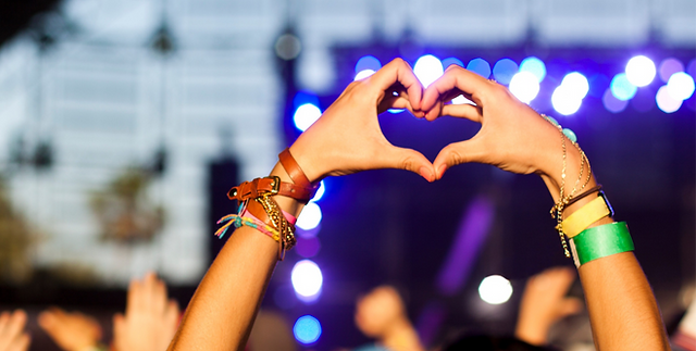 Raised hands with festival wristbands forming a heart shape in front of a glowing blue and purple concert stage Raised hands with festival wristbands forming a heart shape in front of a glowing blue and purple concert stage