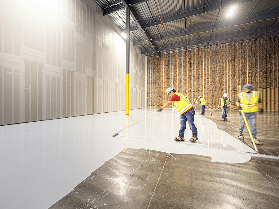Crew in safety vests spreading white coating over a concrete floor inside a large steel framed building with drywall and insulated stud walls