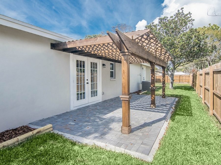 Stained wood pergola by Onsite Construction over a gray paver patio beside white stucco walls and French doors