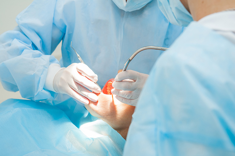 Photo of close up of two people wearing blue surgical gowns and masks with white gloves performing surgery on a person with thier mouth open with a light shining.