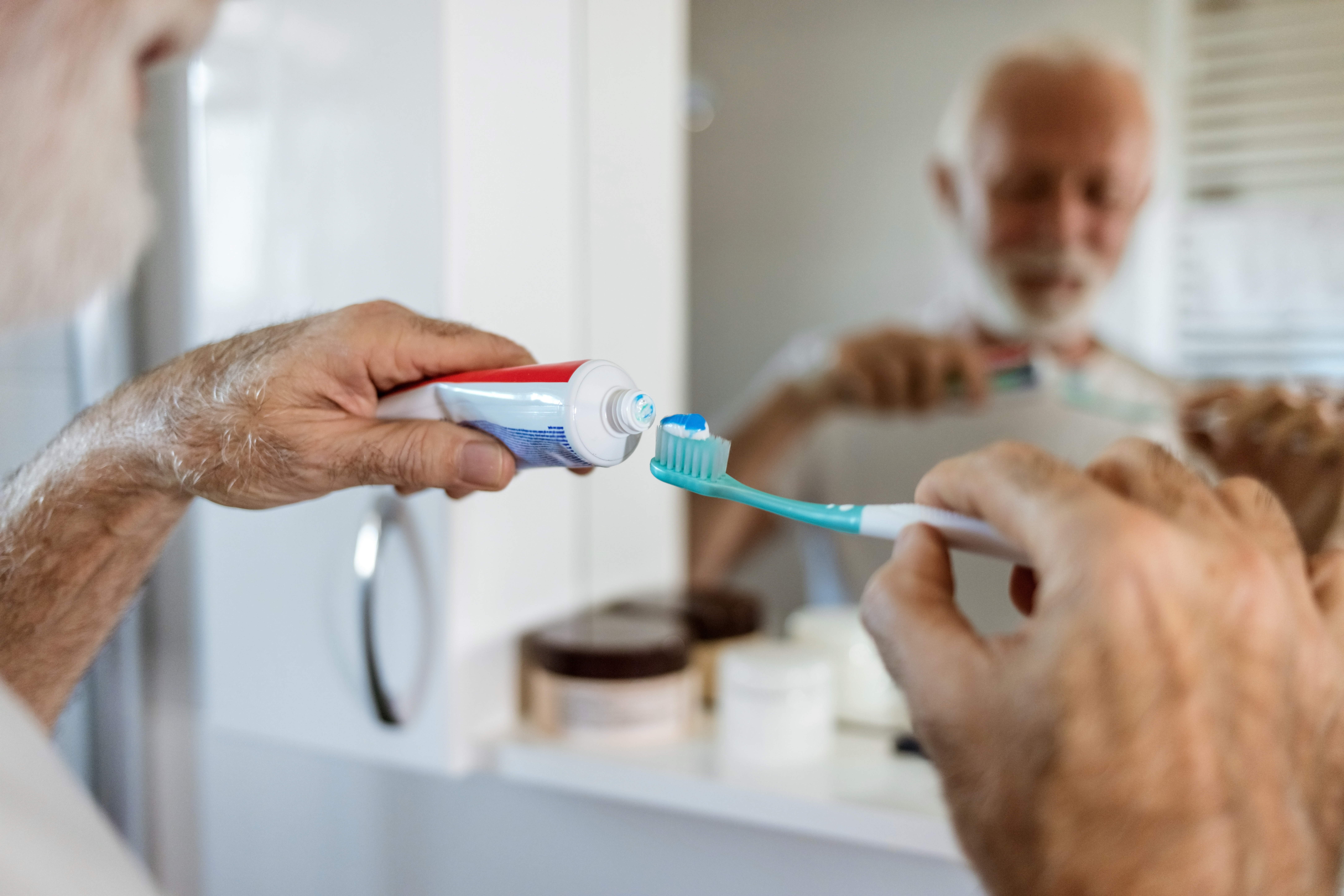 Old man with white goatee holding toothpaste in his left hand squeezing it onto his toothbrush in his right had. He is standing in front of the mirror showing his reflect in the bathroom. 