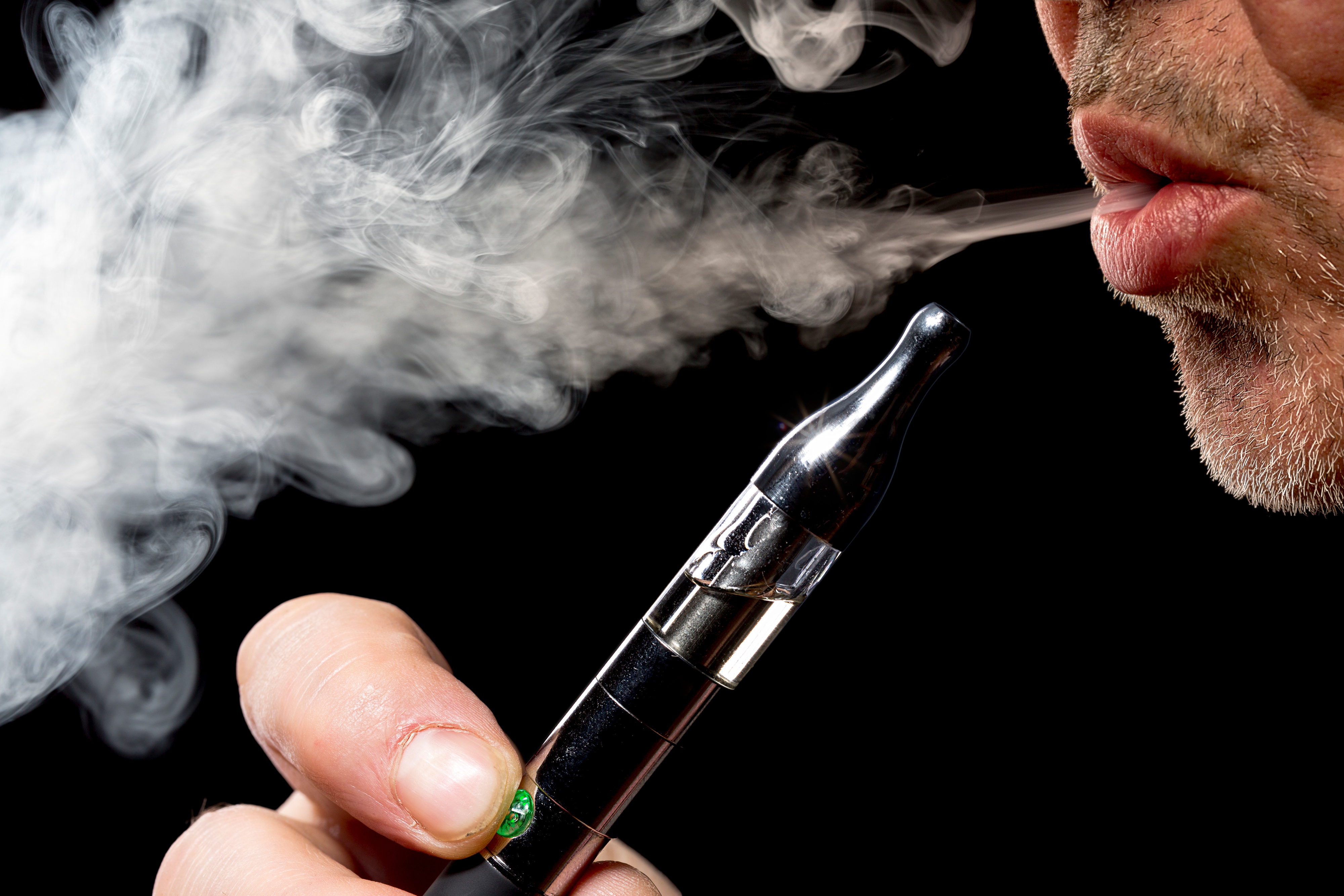 Close-up of the bottom profile of a man's mouth with salt and paper scruffy facial hair using an e-cigarette, blowing smoke, and holding a black vape pen in his right hand. Highlighting the risk of smoking on gum disease.