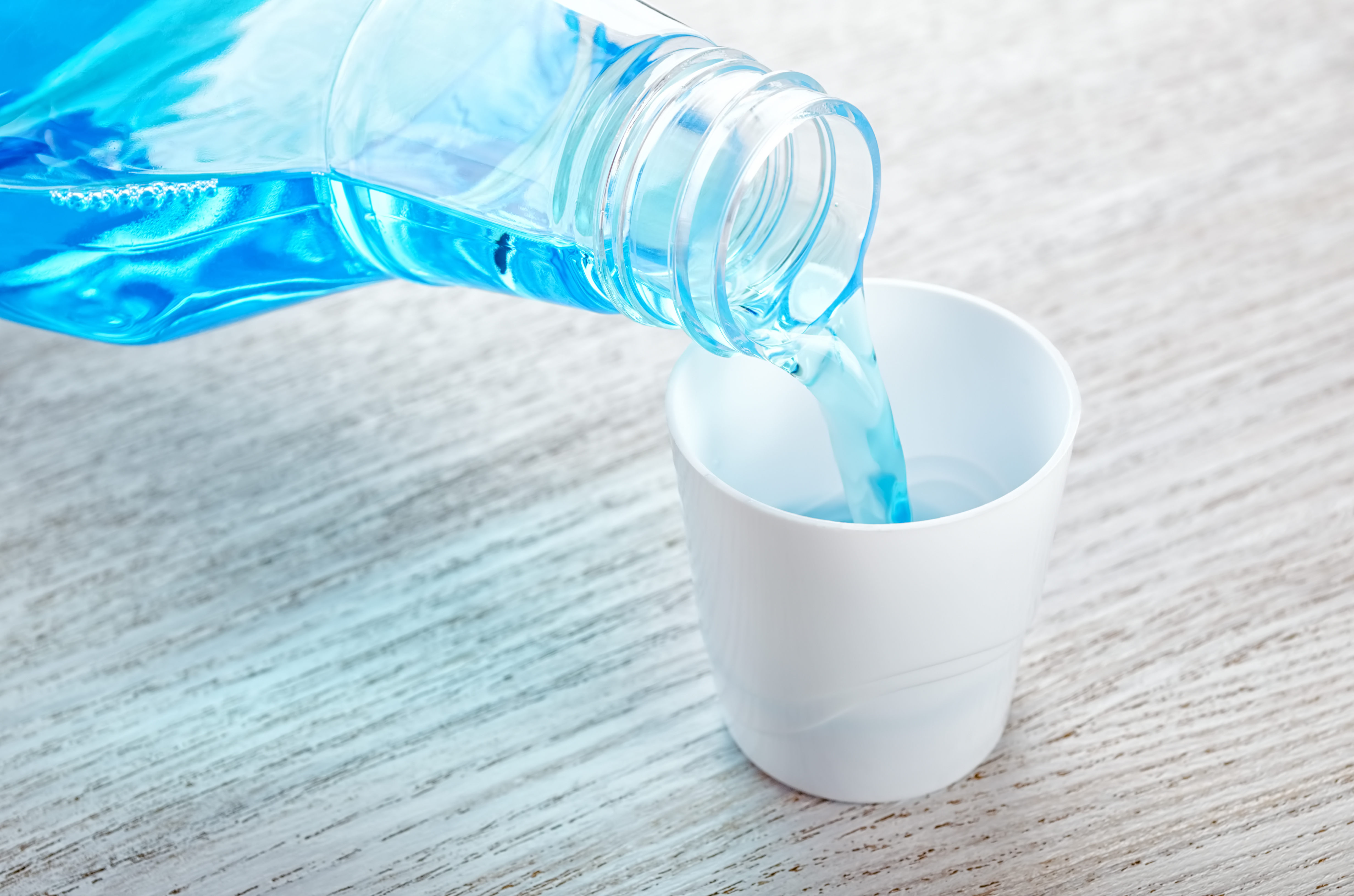 Weathered grey wood surface with a tight shot of a bottle of blue mouth wash being poured into the white cap from the mouthwash