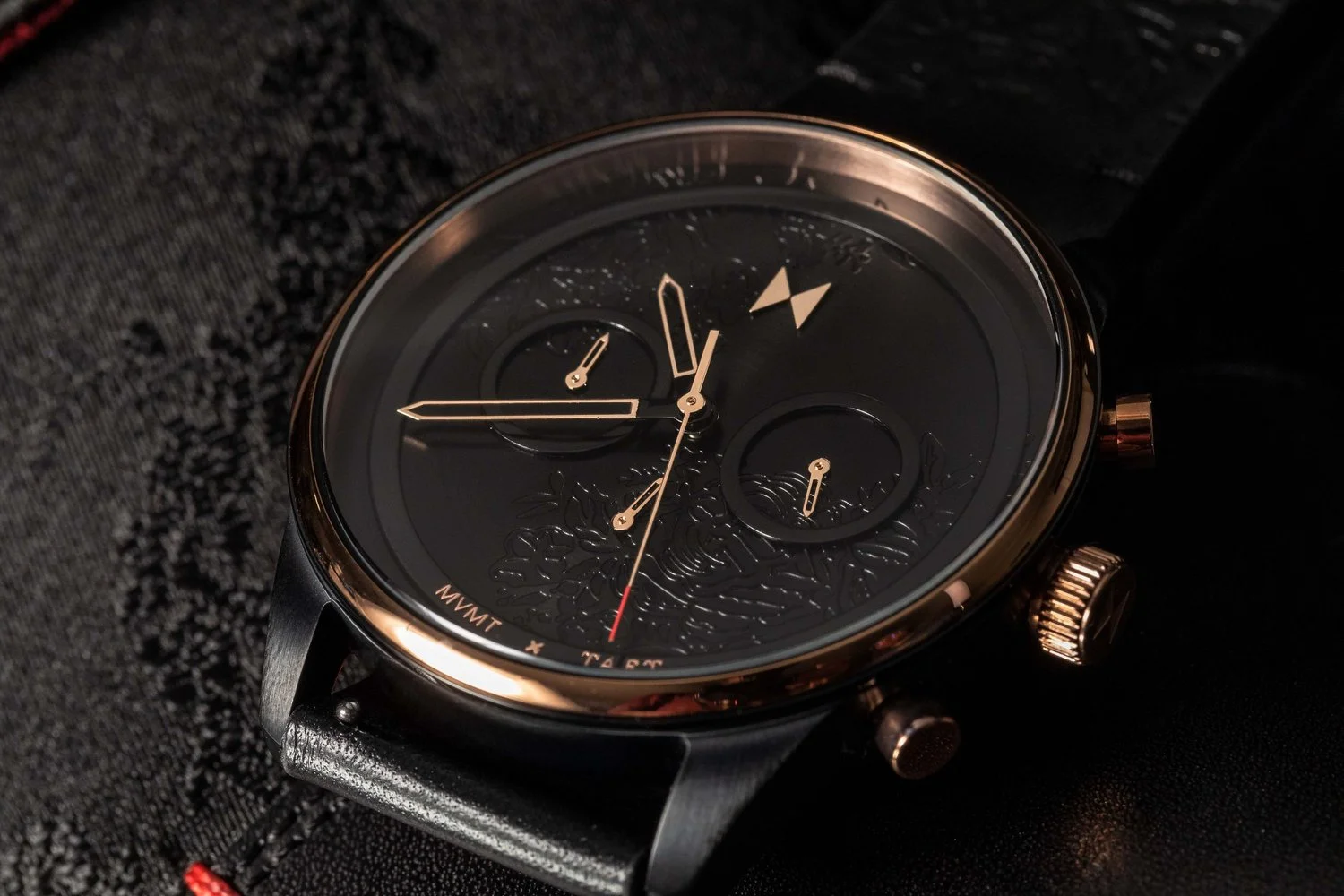 Close-up product photograph of a black MVMT x TAT watch with warm metallic details