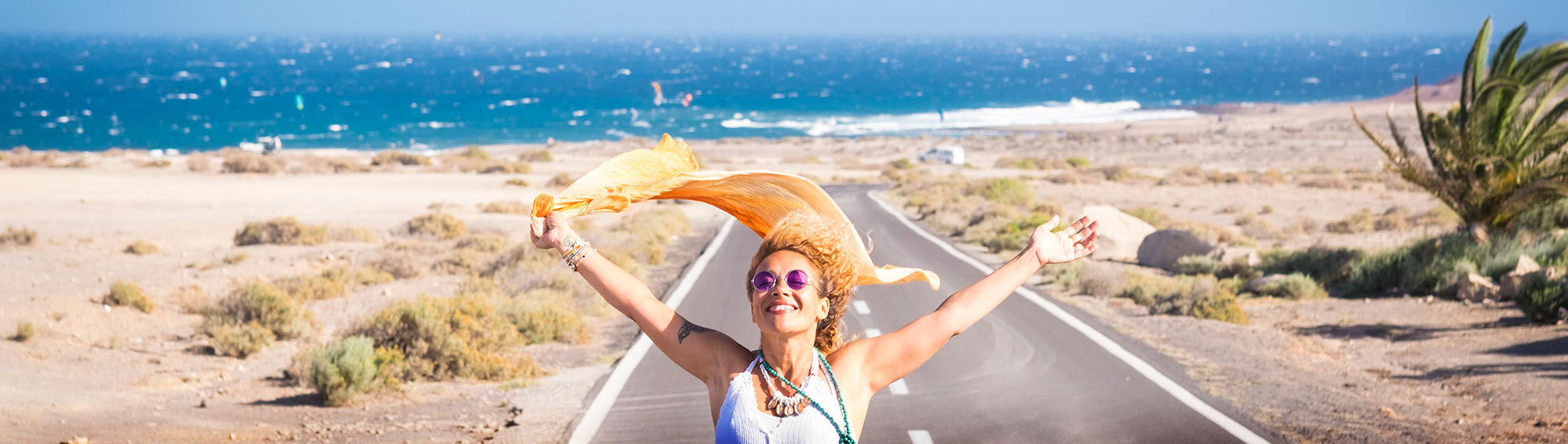 Photo of girl with arms up and back, the wind blowing a scarf she’s holding, standing on a road– the ocean visible in the distance