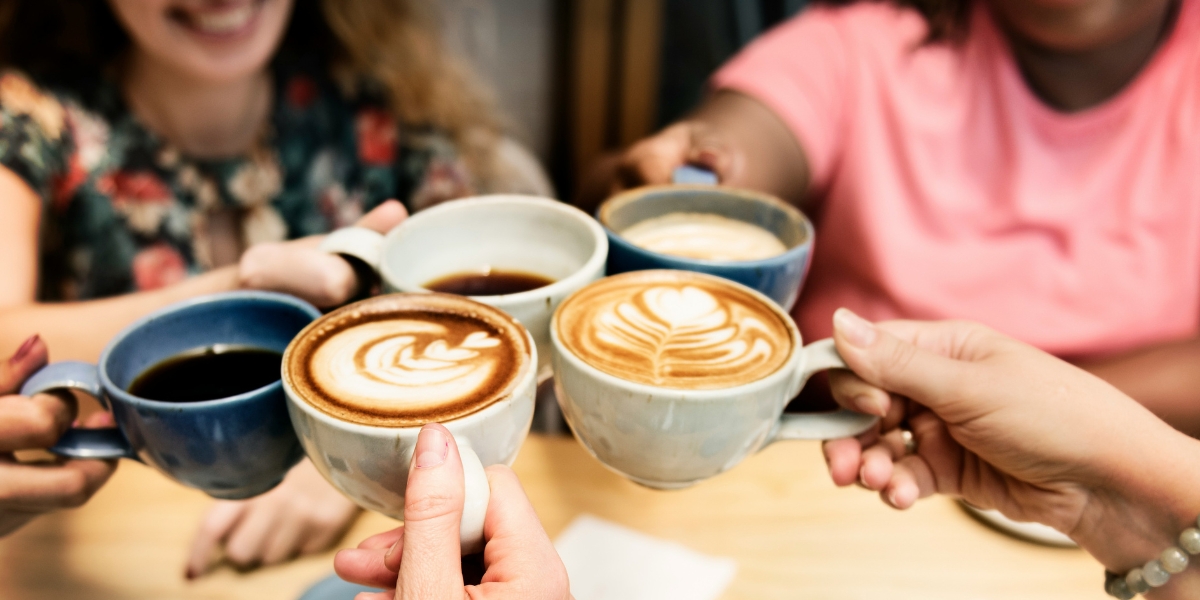 A close-up shot of a group of friends clinking five coffee mugs together in a toast over a wooden table, featuring lattes with decorative foam art and black coffee.