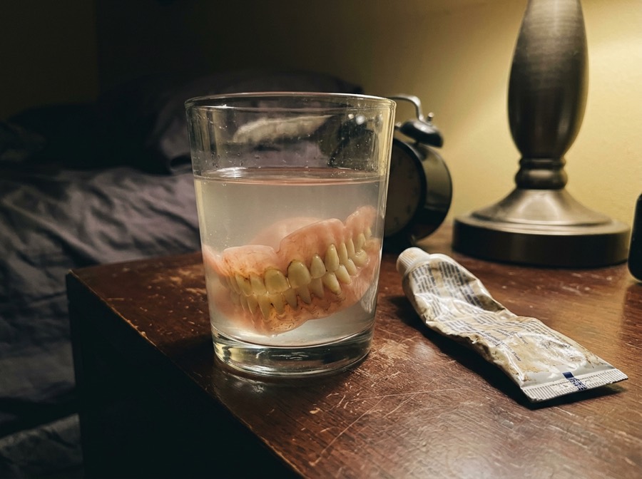 A worn denture soaking in a cloudy glass of water on a nightstand beside a squeezed tube of adhesive