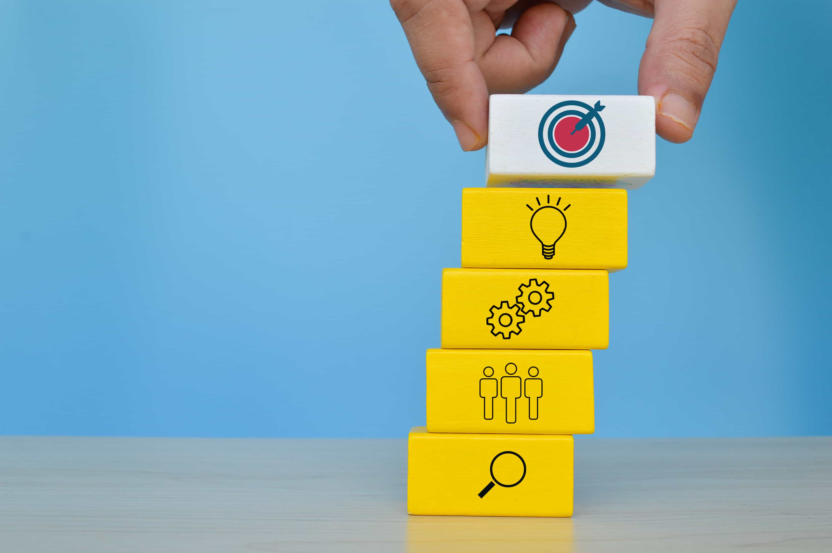 A hand placing a block with a target icon onto a stack of yellow blocks representing business strategy planning in San Diego, CA. A hand placing a block with a target icon onto a stack of yellow blocks representing business strategy planning in San Diego, CA.