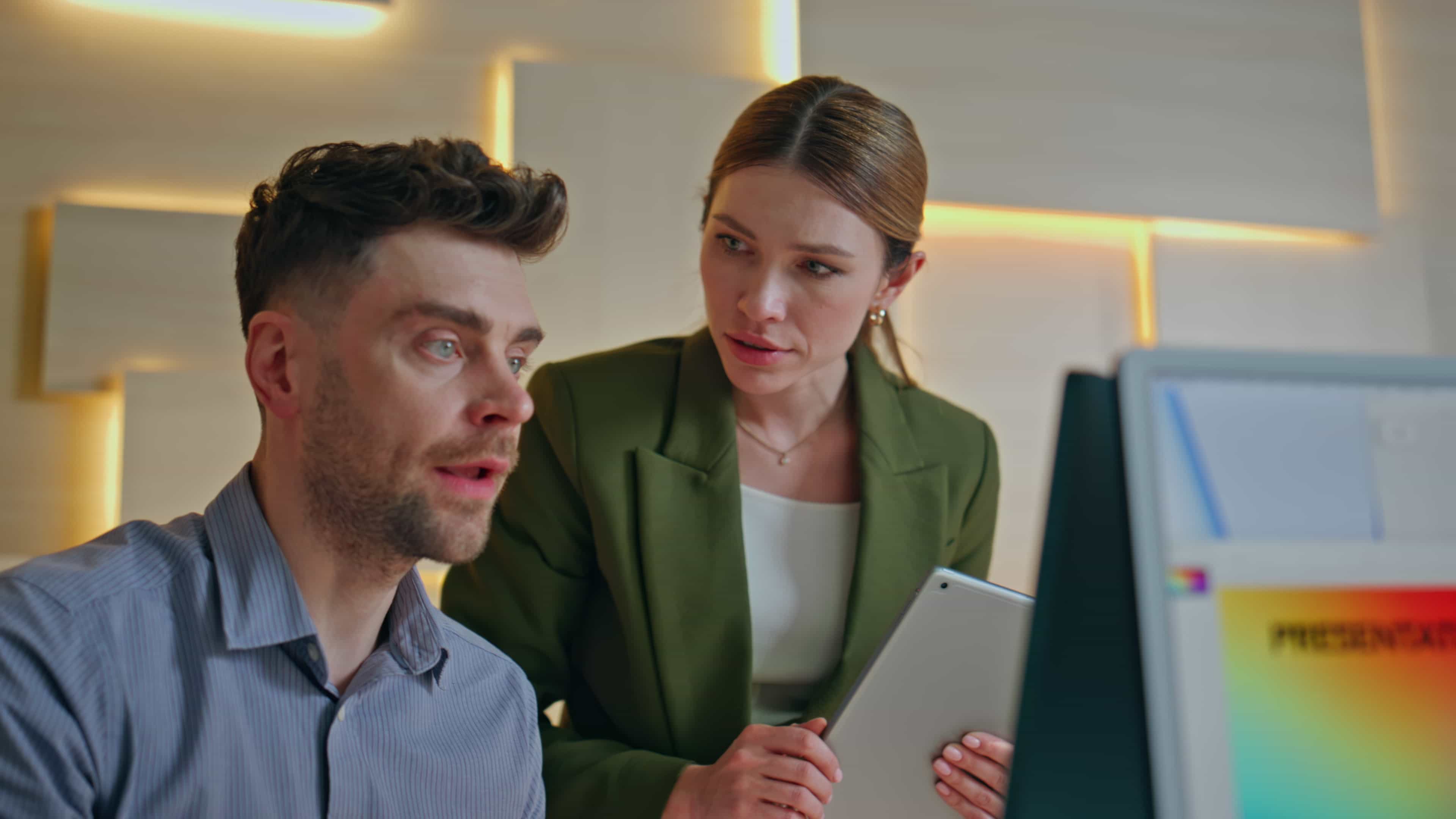 A female executive with a tablet providing corporate leadership training and business consulting services to a male manager looking at a computer screen in a New York office.
