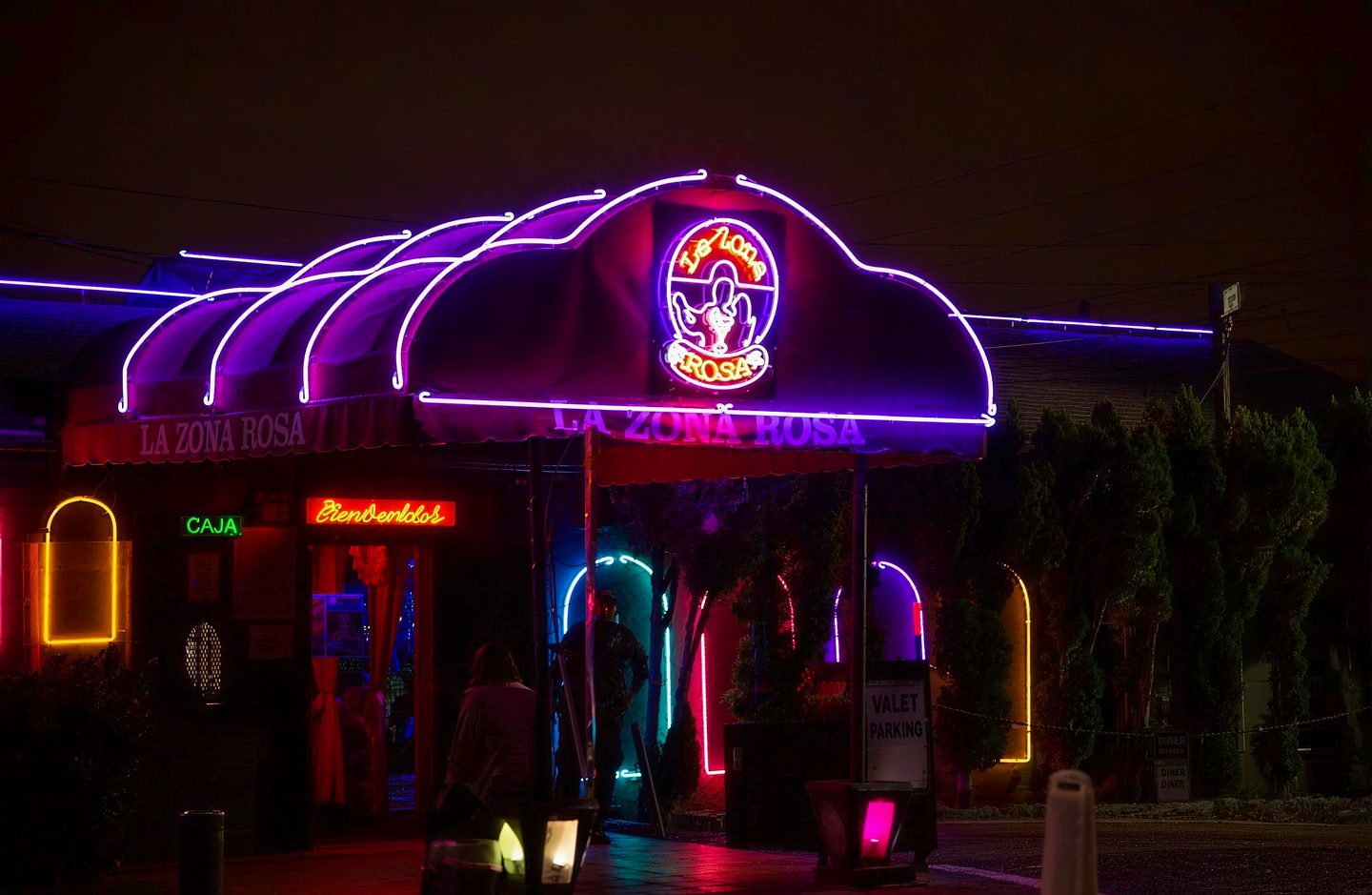 La Zona Rosa's curved awning outlined in violet and pink neon at night, with the crest sign lit above the entrance