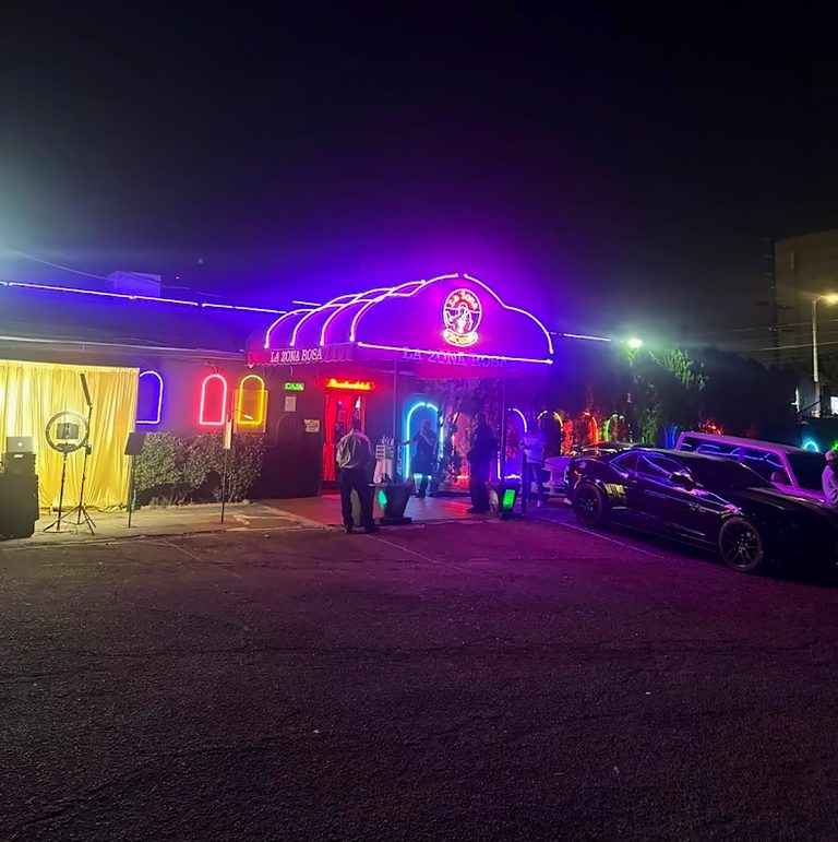 Wide view of La Zona Rosa's neon façade and parking lot at night
