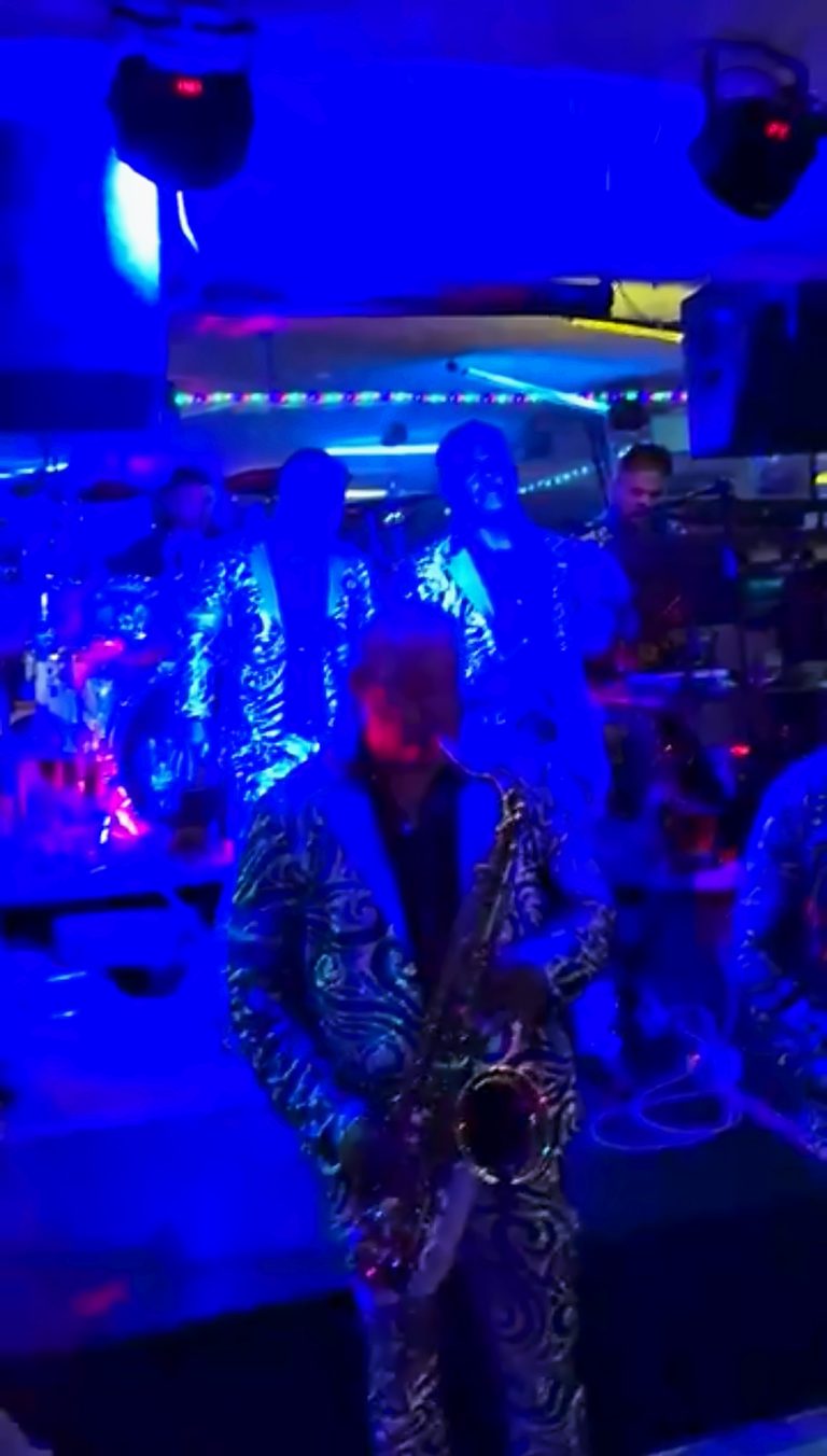 A saxophonist in a sequined jacket playing under blue stage light with the band behind him