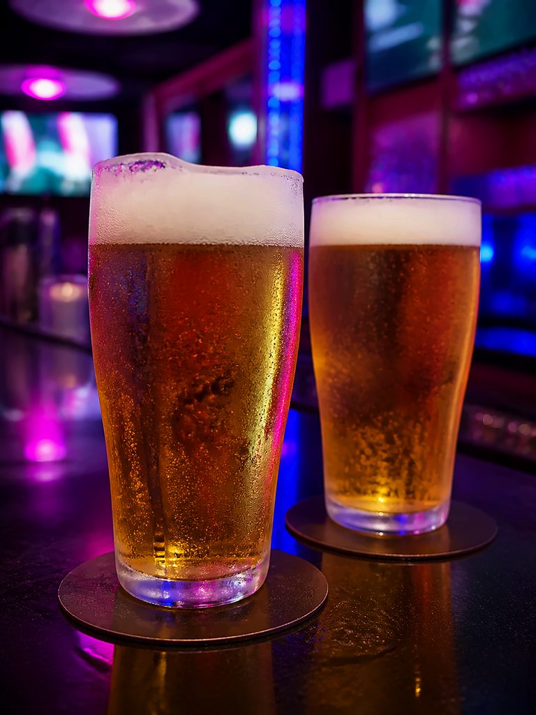 Two freshly poured golden draft beers with foamy heads, bubbles, and condensation on a neon-lit bar