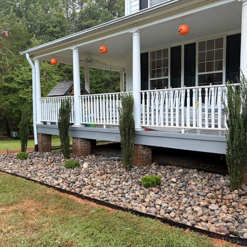 New river-rock landscape beds with junipers and a Japanese maple along a white farmhouse porch installed by Keller Lawns