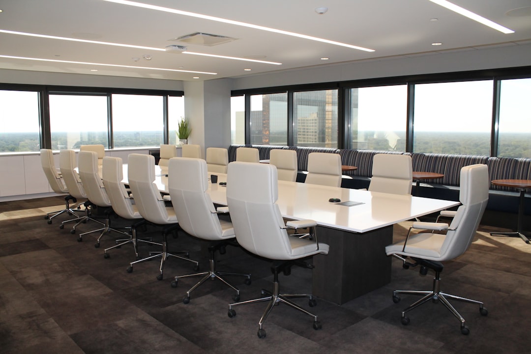 Empty boardroom with long white table, white leather chairs and windows overlooking a city skyline and trees.