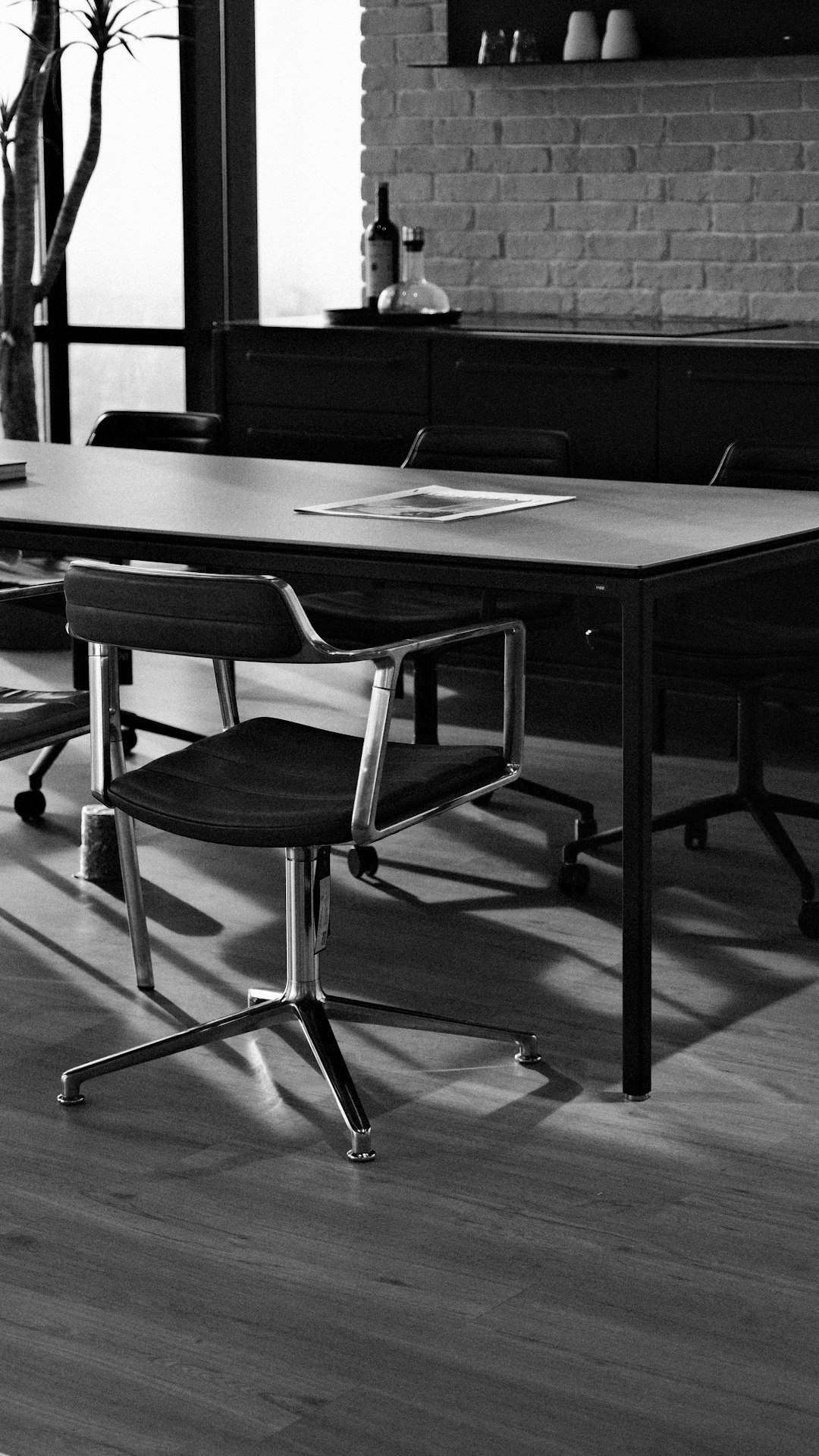 Black-and-white view of an empty conference table with swivel chairs and a document, brick wall behind.