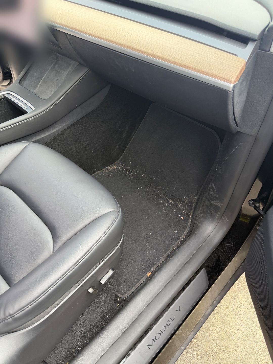 Front passenger footwell of a black Tesla Model Y before detailing, with sand and crumbs across the floor mat