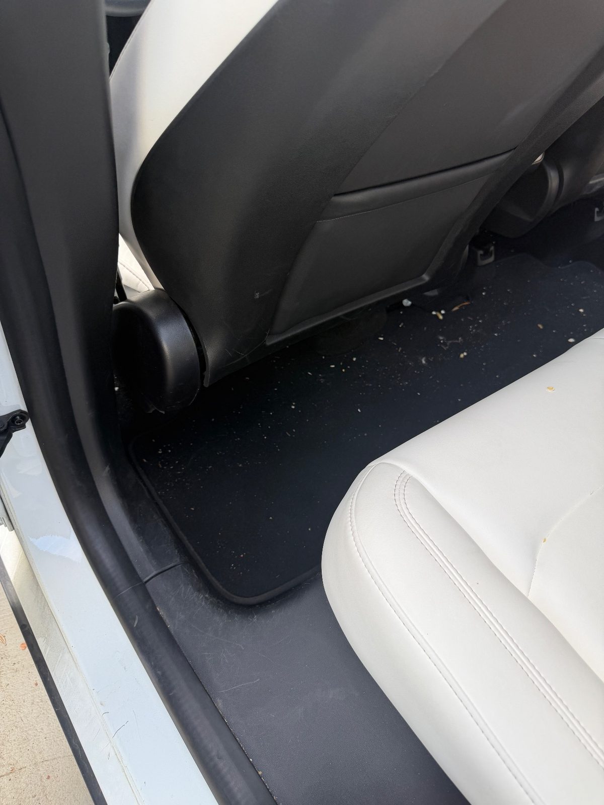 Rear footwell of a white Tesla Model Y before detailing, with crumbs and debris scattered on the carpet mat