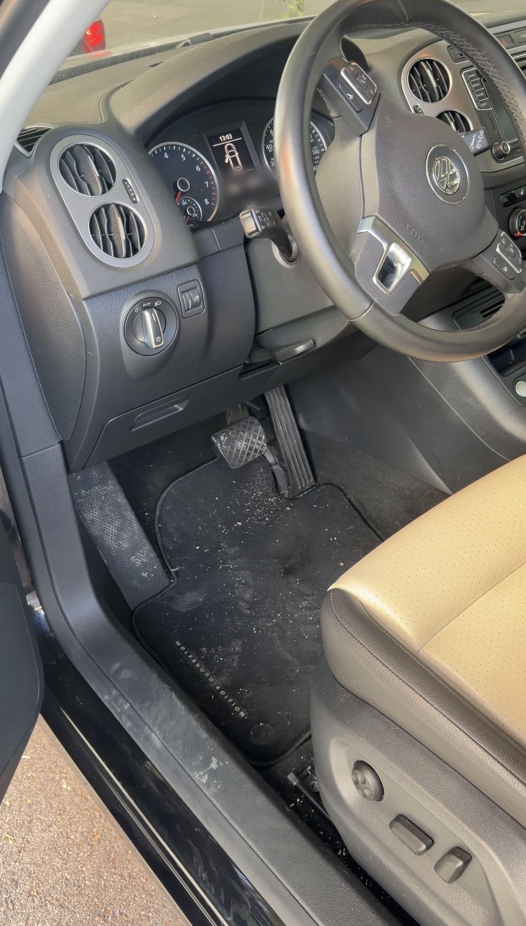 Driver's footwell of a black VW Tiguan Wolfsburg Edition before detailing, loose crumbs and grit scattered across the black carpet floor mat.