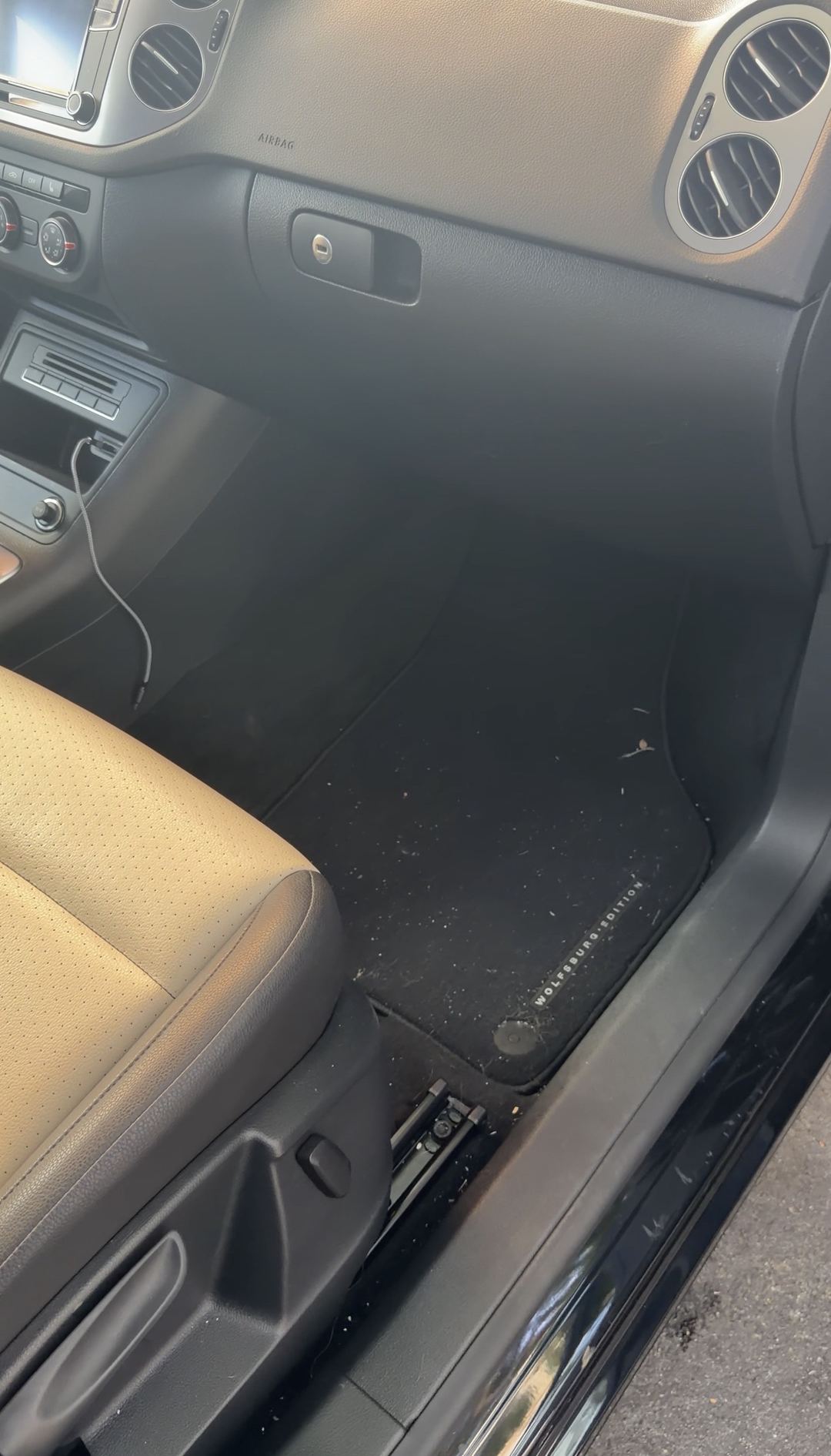 Front passenger footwell of a black VW Tiguan before detailing, crumbs and debris on the Wolfsburg Edition floor mat.