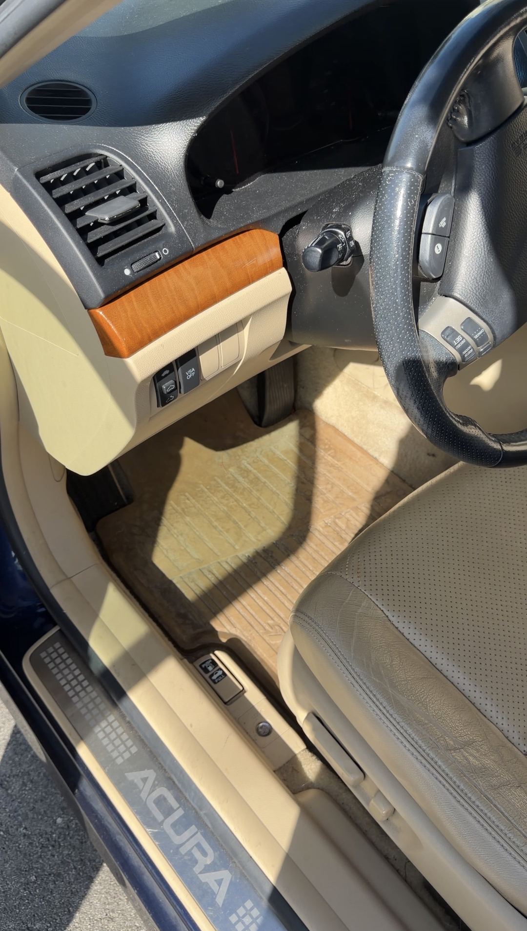 Driver's footwell and lower dashboard of an Acura sedan before detailing, with a dust film on the steering column and a dirt-filmed rubber floor mat.