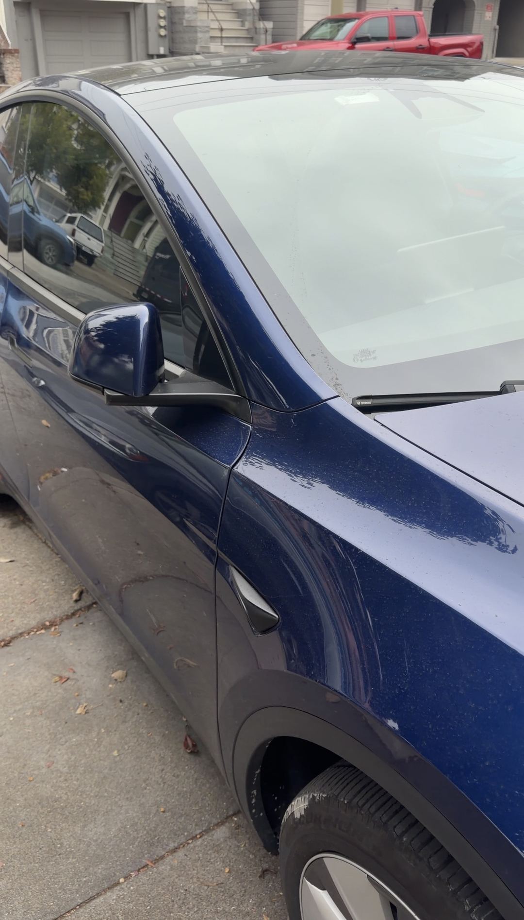 Front corner of a blue Tesla Model Y before washing, with road film on the tyre and water spots on the paint.