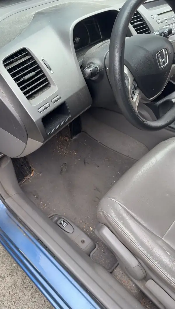 Driver footwell of a Honda Civic before cleaning, with leaf litter piled by the dead pedal and dirt ground into the carpet.