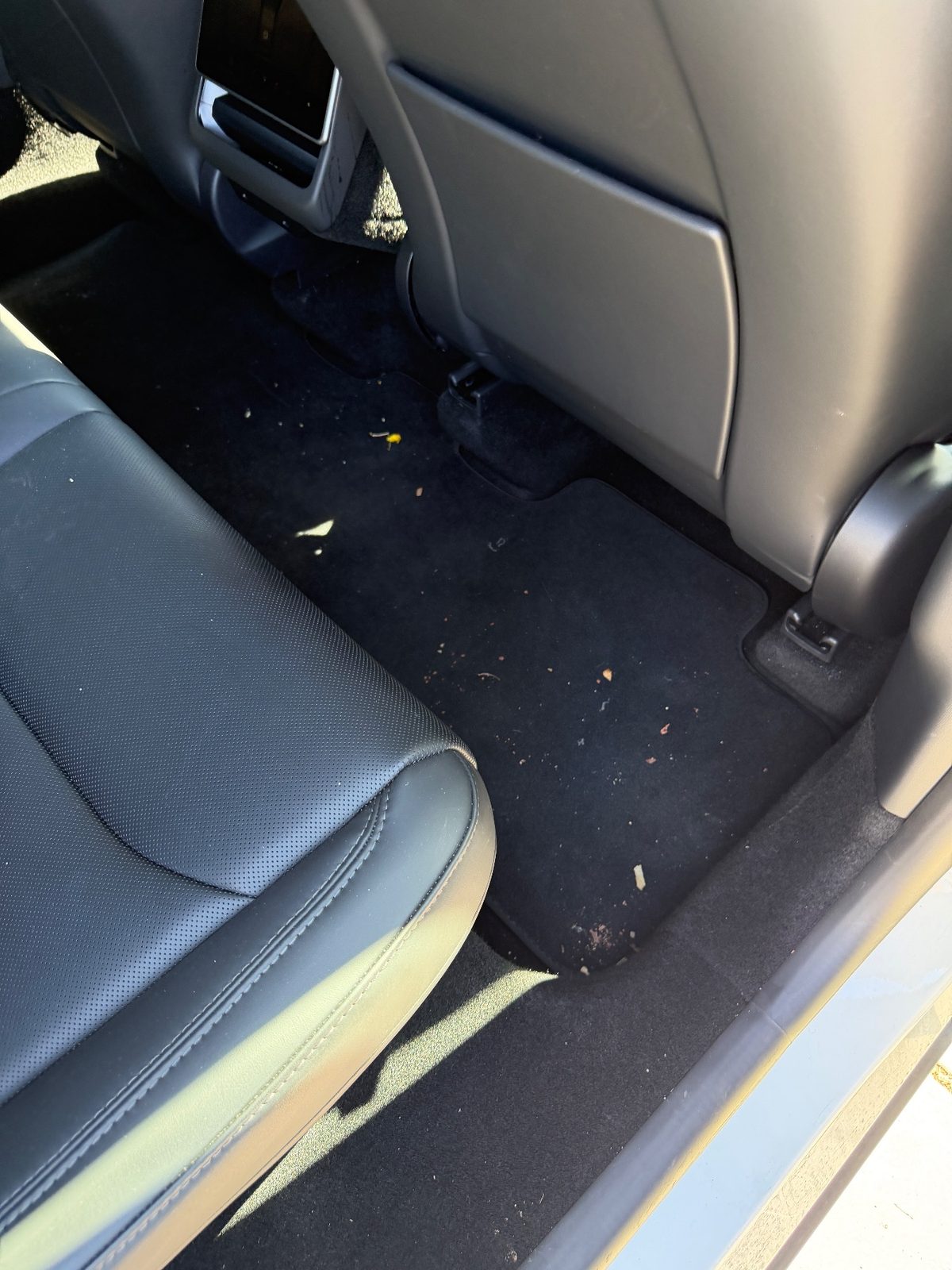 Rear footwell of a white Tesla Model Y before vacuuming, crumbs and leaf bits on the black mat