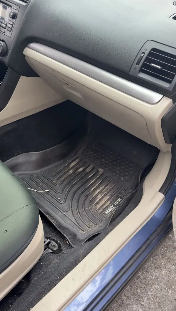 Front passenger footwell of a blue Subaru before cleaning, sandy dirt packed into the grooves of the black rubber floor liner.
